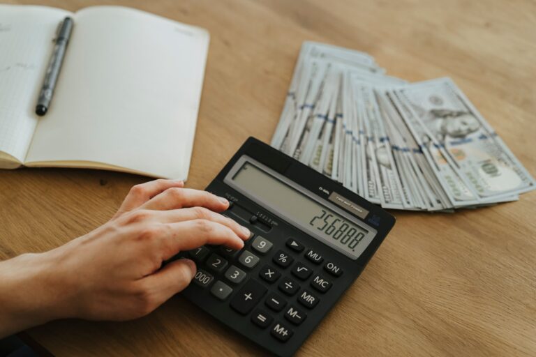 Hand calculating numbers on a calculator next to a notebook and cash on a wood desk, representing running the numbers on a real estate investment.