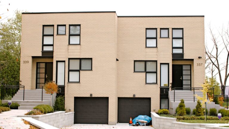 A two-unit modern brick duplex with two front doors, two garages, and a shared driveway, photographed from the street on an autumn day.