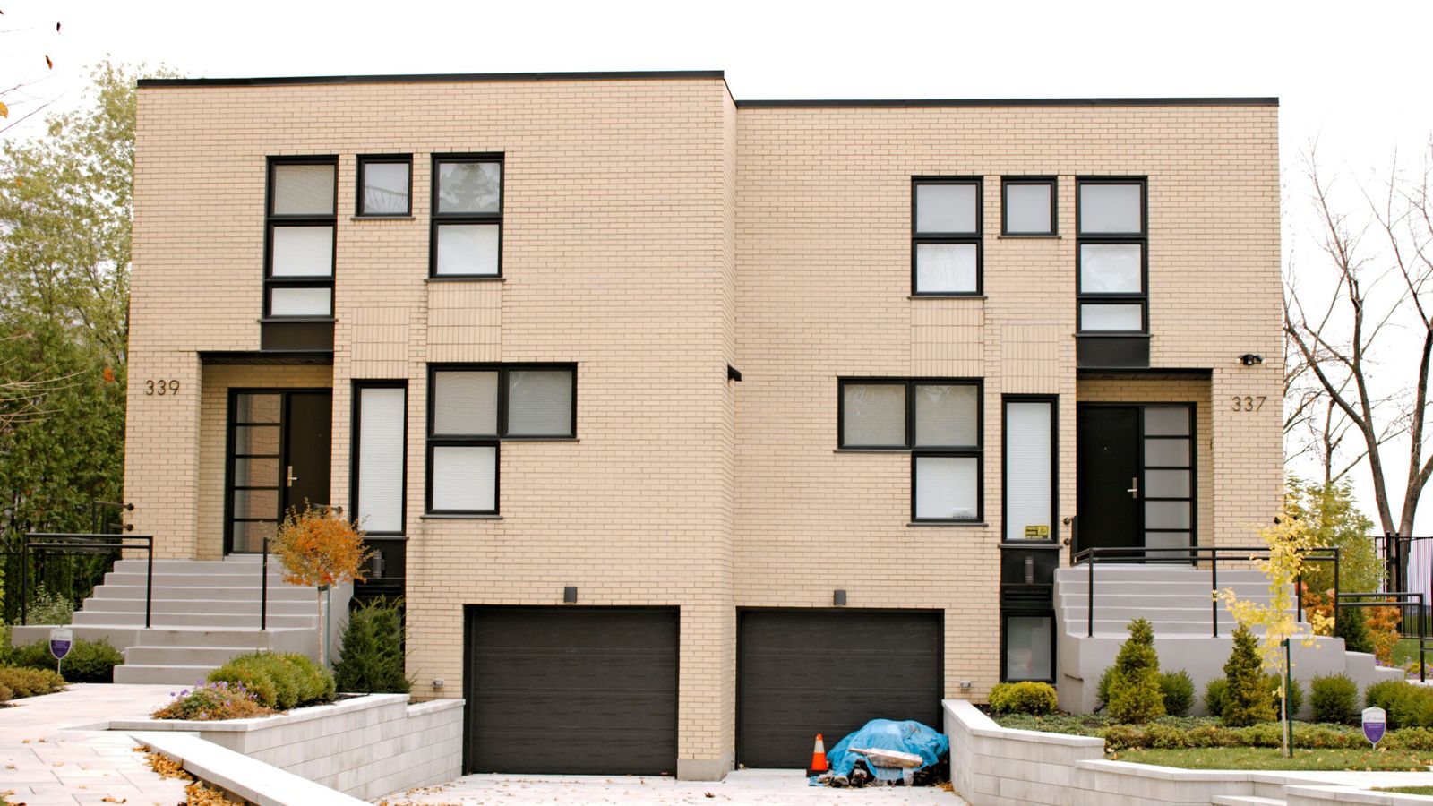 A two-unit modern brick duplex with two front doors, two garages, and a shared driveway, photographed from the street on an autumn day.