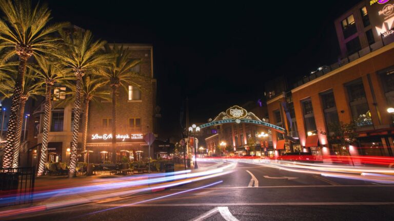 The Gaslamp Quarter entrance arch in downtown San Diego at night, lit storefronts and palm trees along the street with car light trails streaking across the foreground.